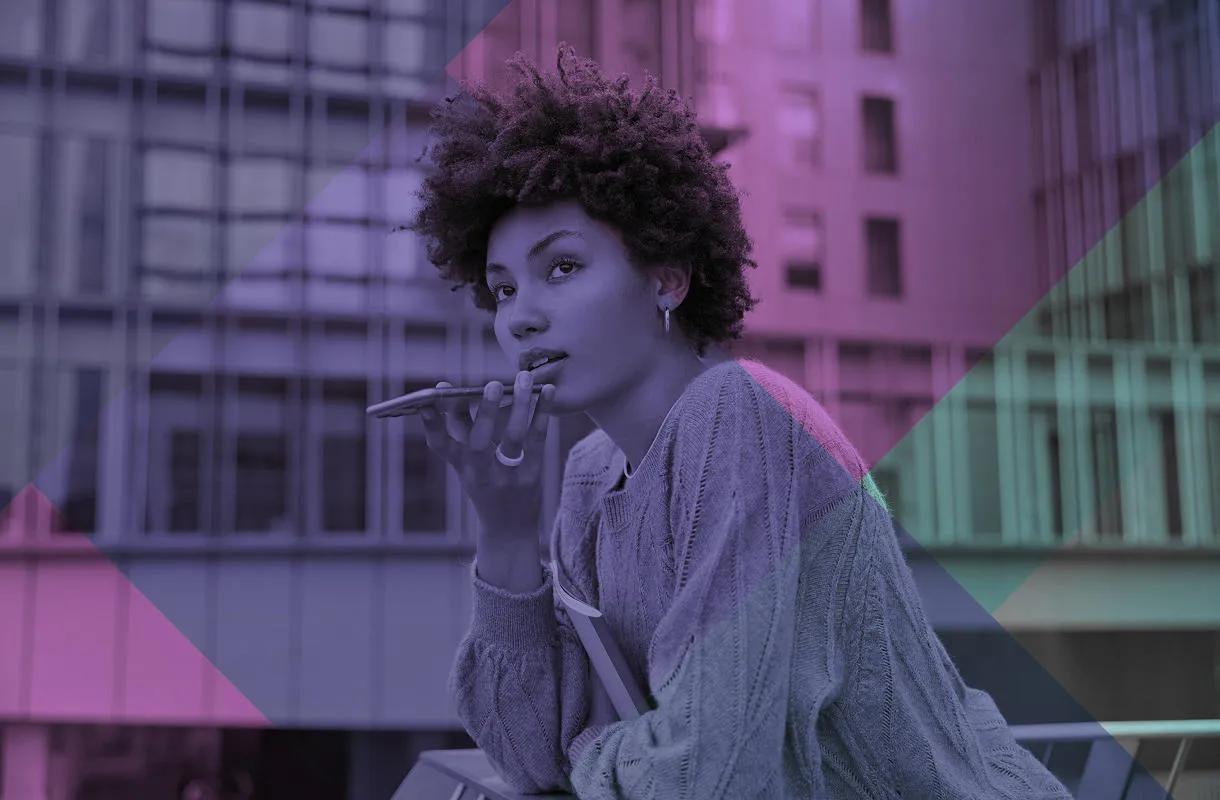 A young woman standing on a balcony or roof terrace in a city and speaking into the bottom of a mobile phone.